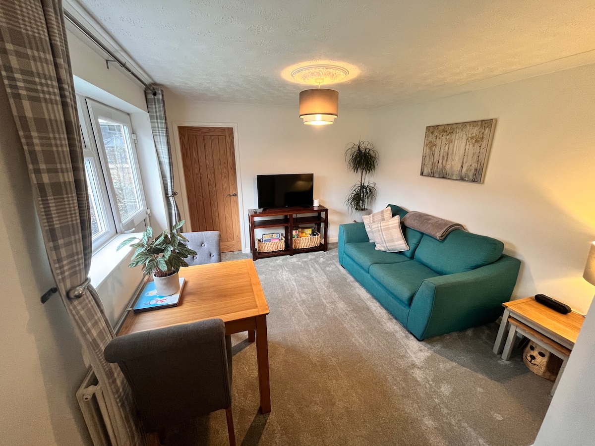 The living room features a bright and spacious layout with a comfortable sofa and a small wooden table. A television is positioned on a stand, with shelves underneath offering storage. Natural light filters in through the windows, complemented by soft lighting fixtures.