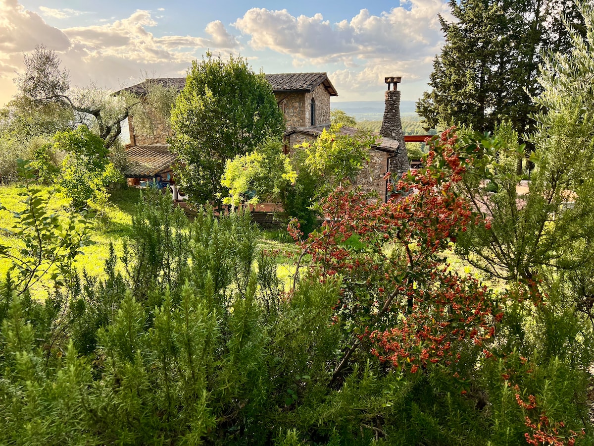 The farmhouse is nestled among lush greenery and colorful shrubs, showcasing rustic stone architecture. A scenic landscape includes trees and flowering bushes, providing a feeling of tranquility. The sky is adorned with scattered clouds, hinting at the beauty of the surrounding countryside.