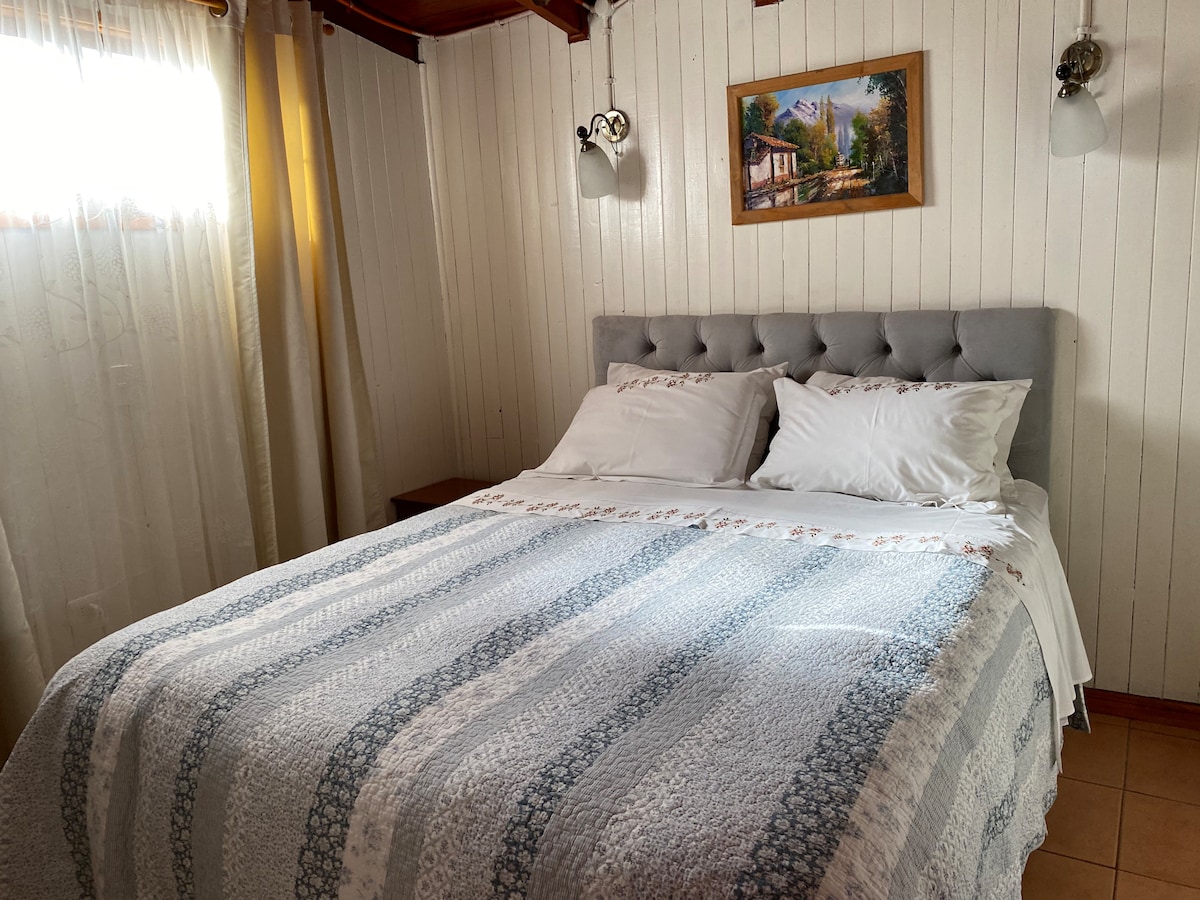 A cozy sleeping area is featured with a tufted grey headboard and white linens. Natural light filters in through a nearby window, casting soft shadows on the light-colored walls, while a framed landscape painting is displayed above the bed.