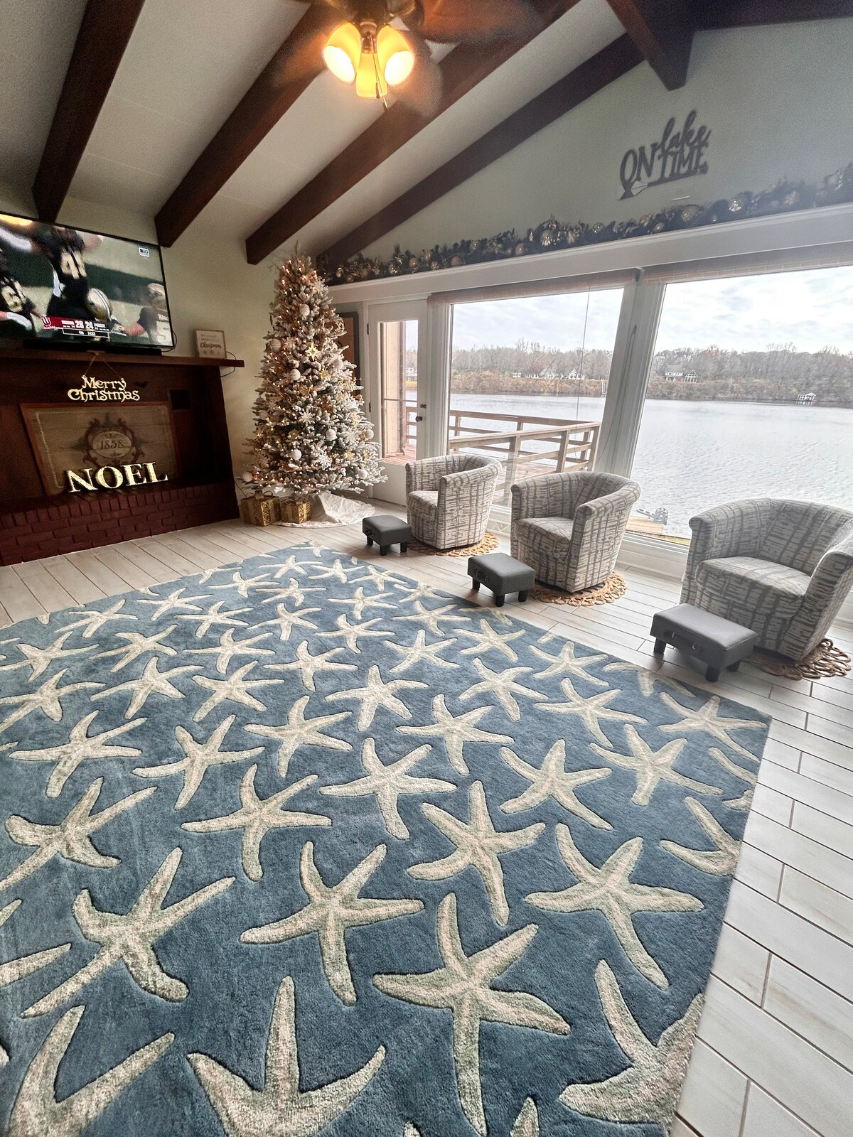 A well-lit living area is adorned with a large area rug featuring starfish patterns. Two wicker chairs are positioned near a large window that overlooks the lake. A decorated Christmas tree and a television are visible near the fireplace, creating a cozy atmosphere.