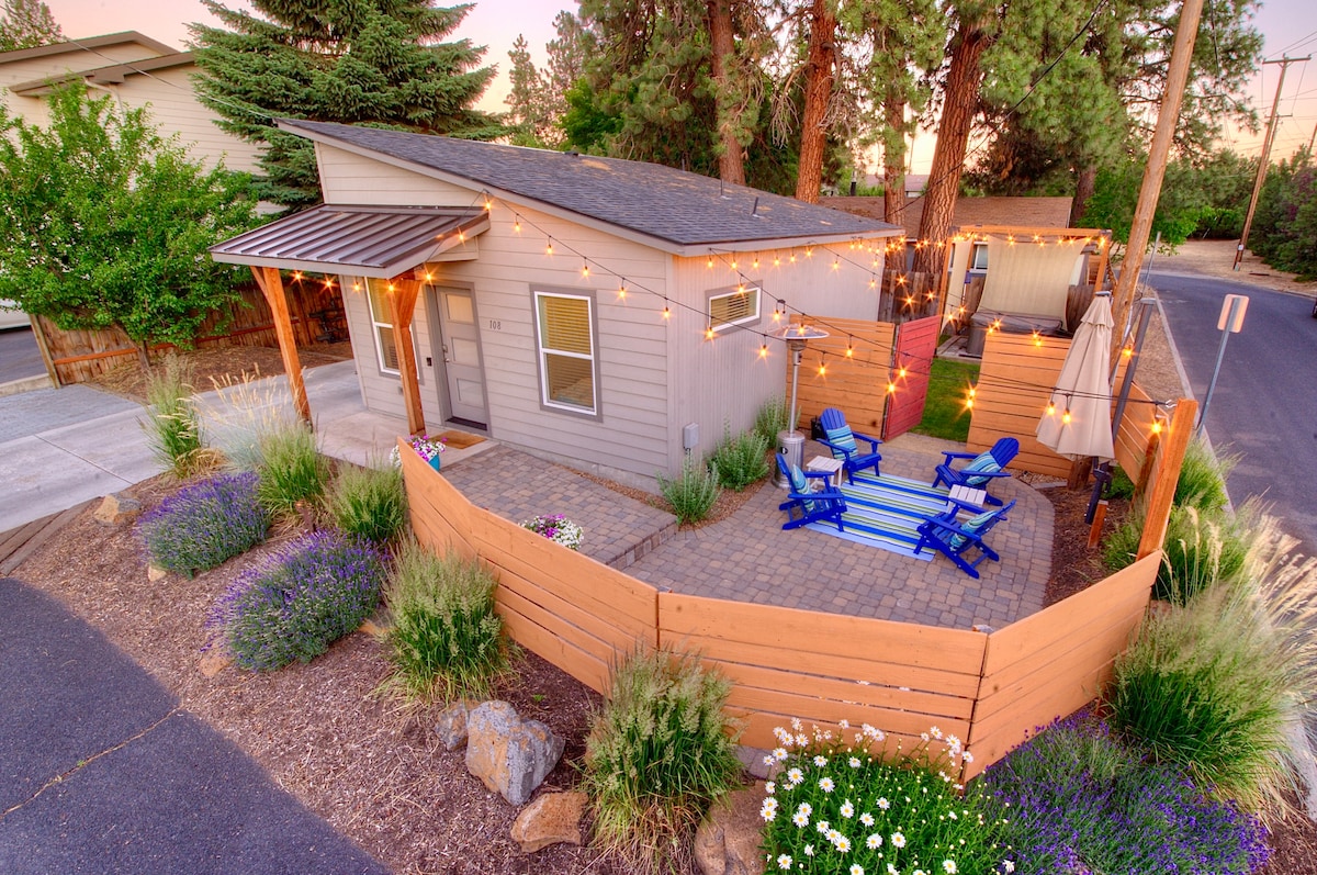 The exterior of the home is surrounded by a wooden fence and adorned with string lights. A cozy seating area with blue chairs is positioned on a striped outdoor rug. Lush plants and flowers enhance the landscape, creating a welcoming entryway.