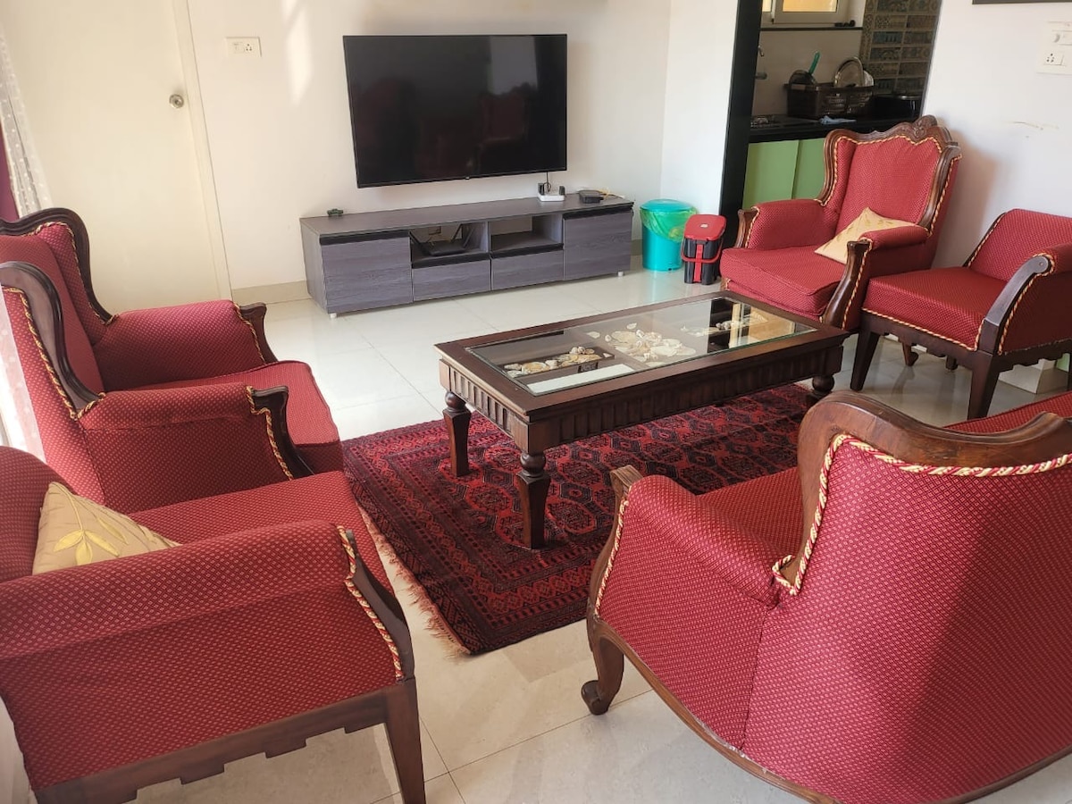 A living area features elegantly arranged traditional seating with red upholstery, complemented by a central wooden coffee table. A decorative rug lays beneath, and a wall-mounted television is visible. The space is well-lit with natural light filtering through nearby windows.