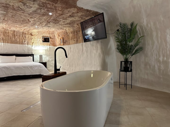 A stylish cave-like room featuring a freestanding bathtub with a sleek black faucet. The space has natural stone walls, a large bed with white linens, and a mounted TV. A potted plant adds a touch of greenery, while soft lighting creates a cozy ambiance.
