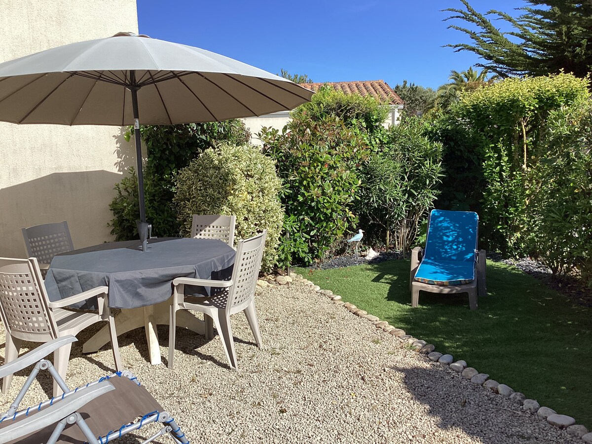 A small garden features a round table covered with a gray tablecloth, surrounded by four beige chairs. A large umbrella offers shade, while a chaise lounge in blue rests on the grass. Lush greenery provides privacy around the space.