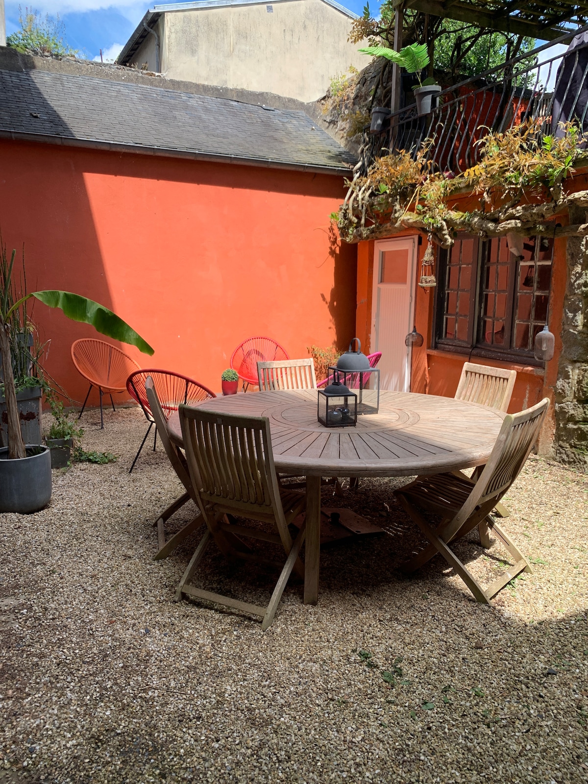 A sunlit courtyard is surrounded by vibrant terracotta walls, featuring a round wooden table set with six chairs. Colorful lounge chairs add a pop of color, while potted plants and hanging flowers enhance the inviting outdoor space.