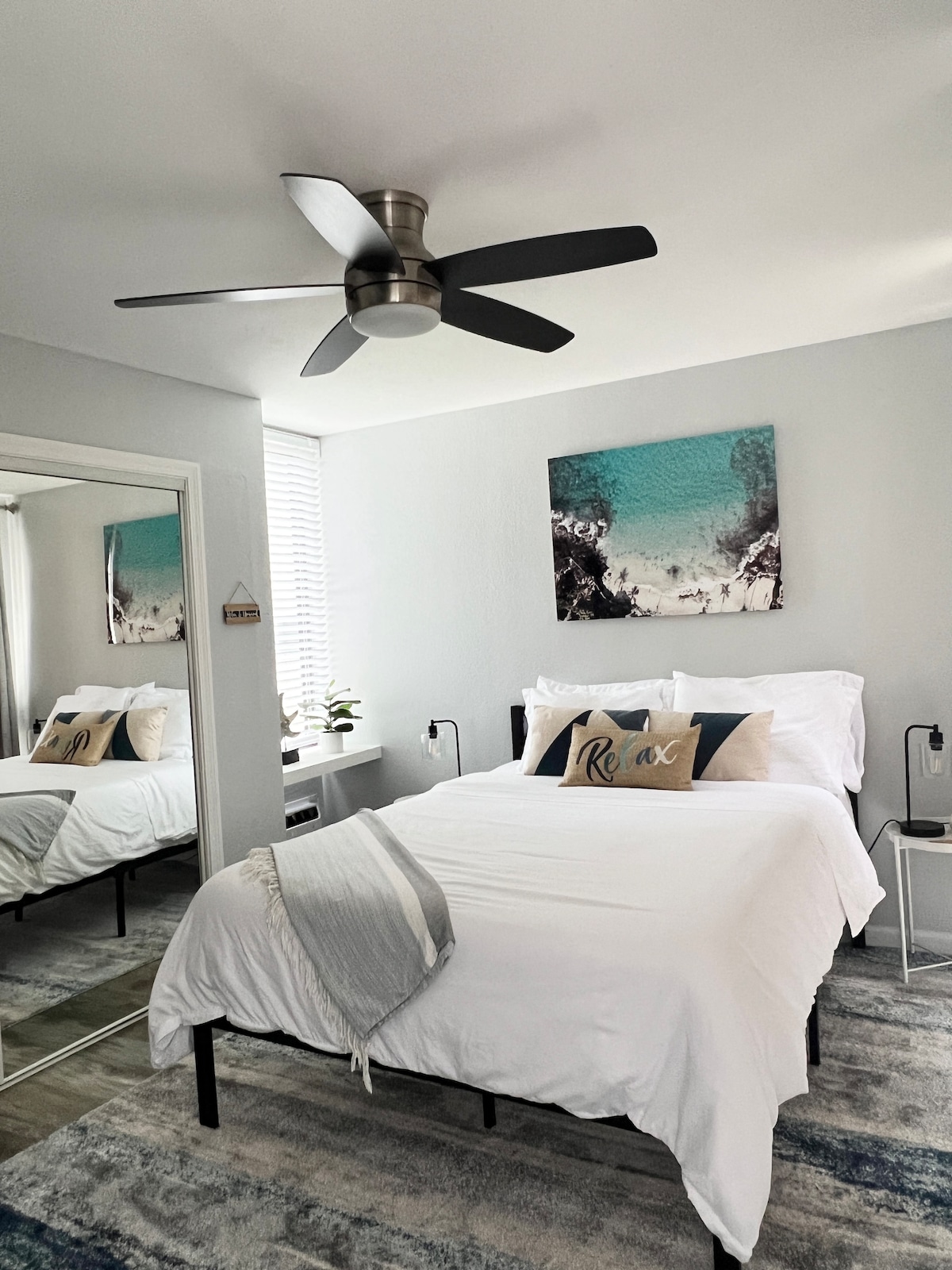 A well-appointed bedroom features a queen size bed dressed in white linens, complemented by decorative pillows. A ceiling fan provides airflow, and a small plant sits by the window. A full-length mirror reflects the room's inviting atmosphere, with art showcasing a serene coastal scene.