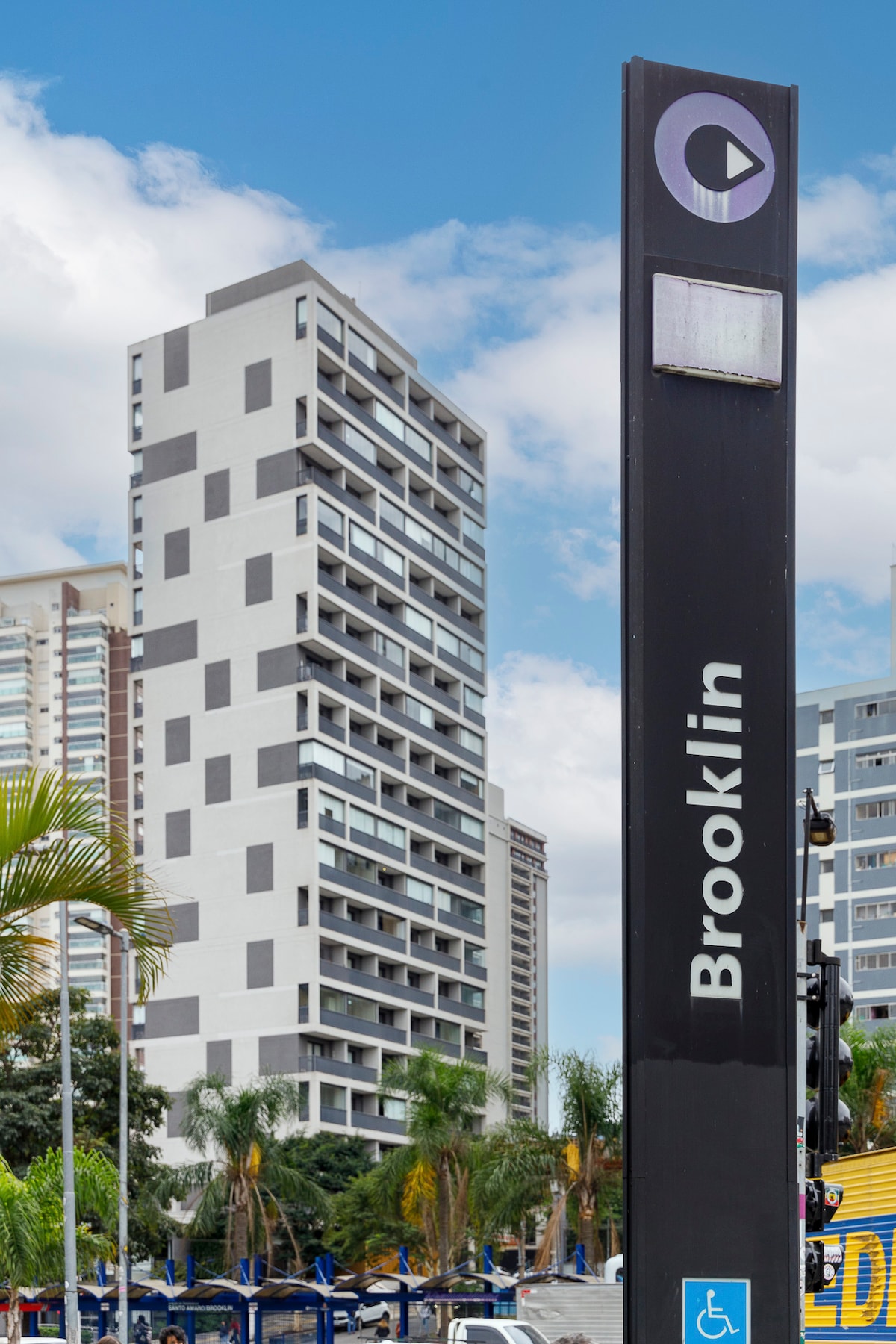 The image features the Brooklin Metro Station sign prominently in the foreground, with a modern high-rise building visible in the background. Lush palm trees and other greenery are present in the area, complementing the urban landscape under a partly cloudy blue sky.