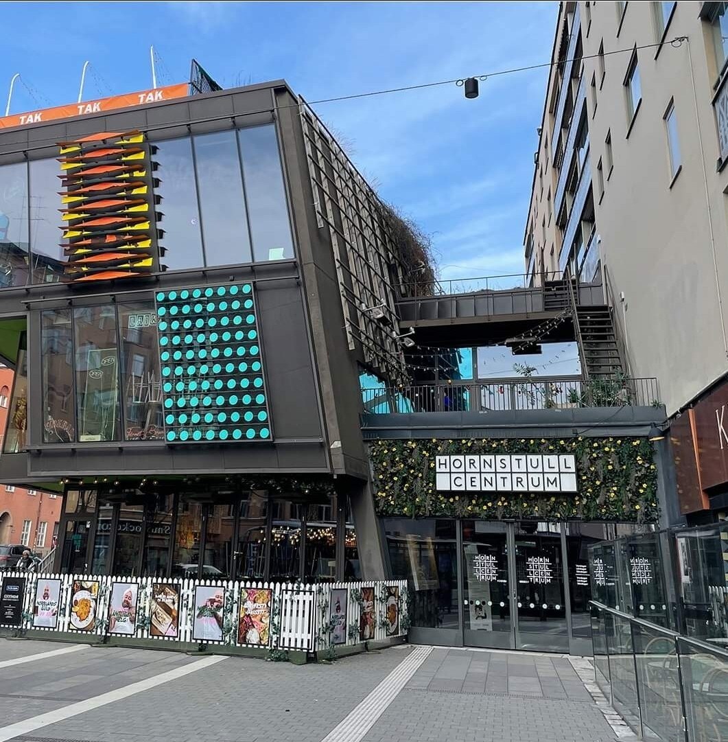 The exterior of Hornstull Centrum features a modern design with multiple levels and colorful facade elements. Lush greenery decorates the upper levels, while the entrance is framed by decorative lights. A wide walkway provides access to the shops, creating a vibrant urban environment.