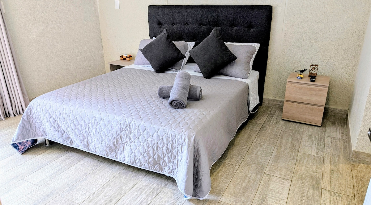 غرفة نوم رئيسية مع سرير بحجم كوين (1.60 م × 2.00 م)، لوح رأس مبطن، وسائد مزخرفة وطاولات بجانب السرير على كل جانب. مثالي لقضاء عطلة مريحة وهادئة.