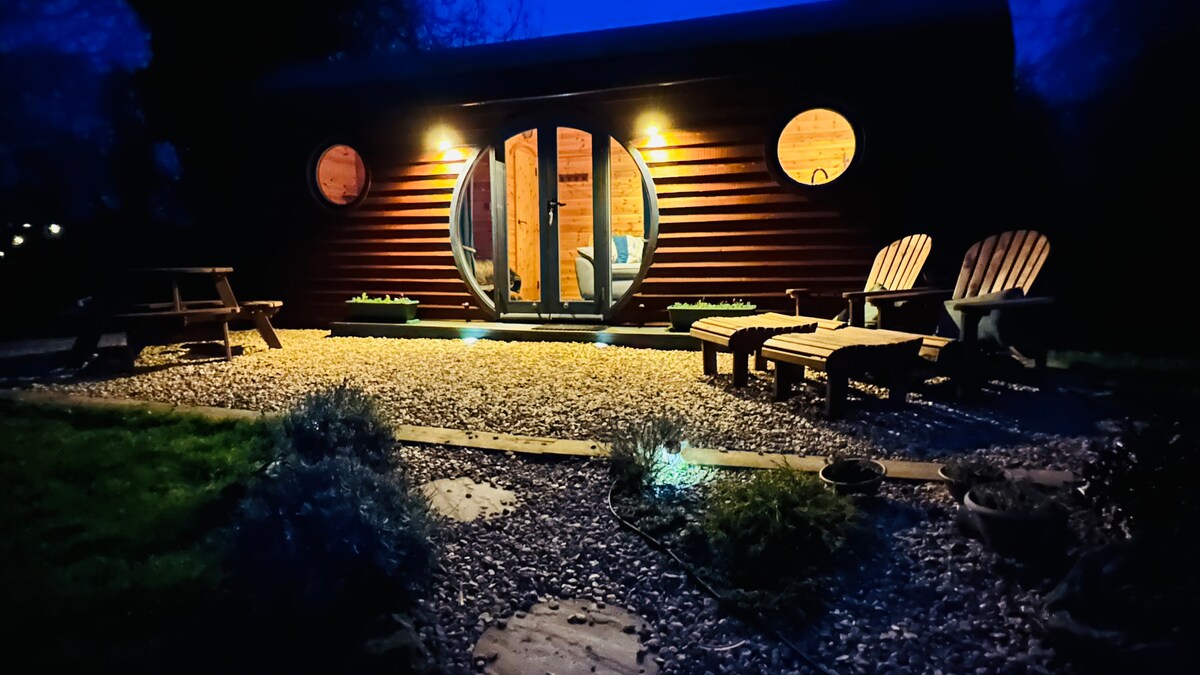 The glamping pod is illuminated by soft lighting at dusk, accentuating its rounded windows and inviting entrance. Wooden loungers and a picnic table provide outdoor seating options, surrounded by a gravel pathway and small greenery, creating a welcoming atmosphere for relaxation.
