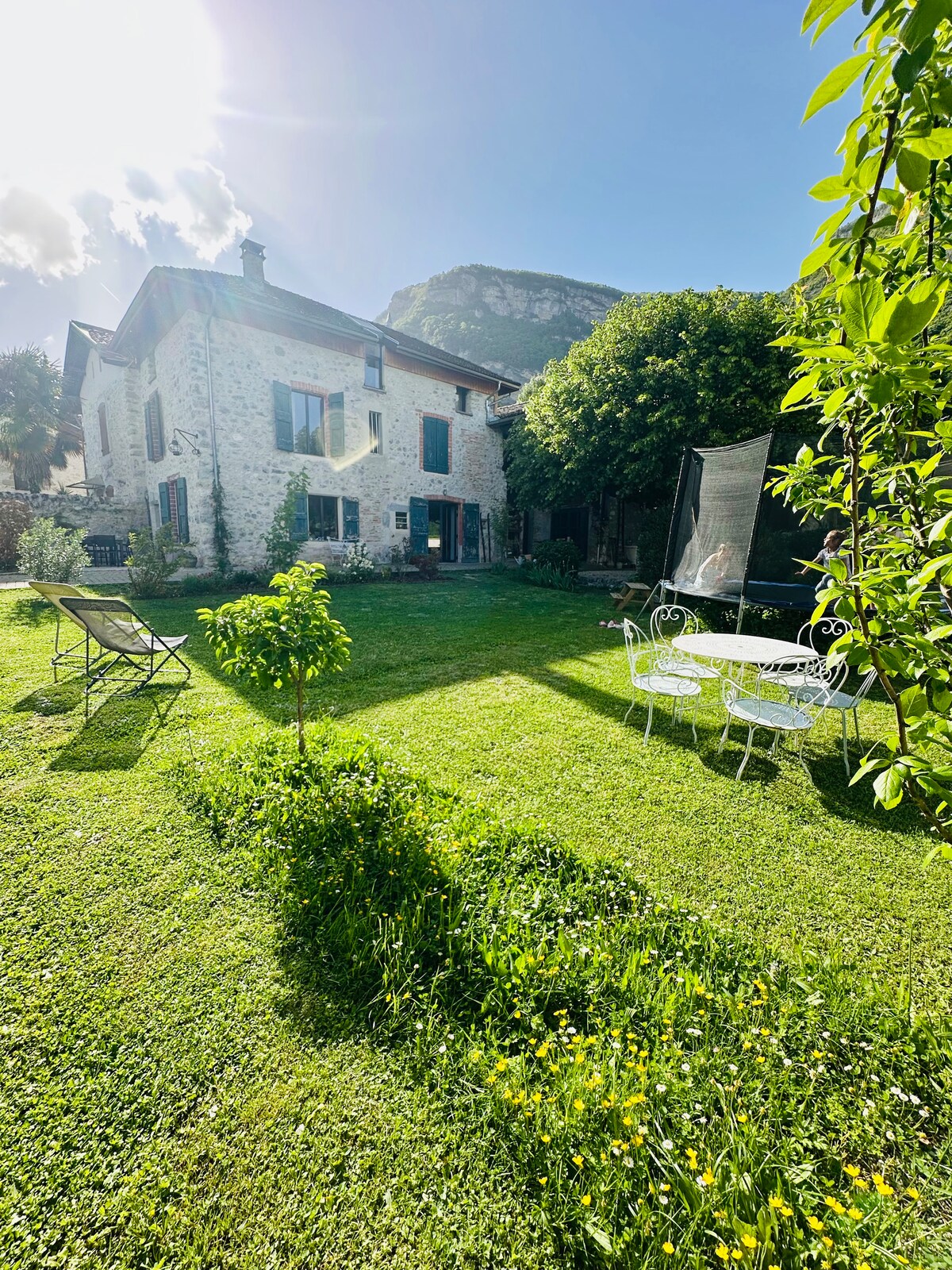 A stone house is set amid lush greenery, with sunlight illuminating the inviting garden. A small tree and colorful flowers add charm to the lawn. White garden furniture is positioned on the grassy area, with a backdrop of mountains creating a serene environment.