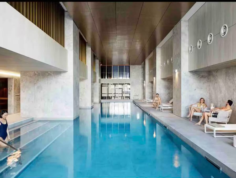 An indoor pool is seen, featuring inviting blue water that reflects the surrounding marble walls. Lounge chairs are positioned along the edges, providing a space for relaxation. Natural light filters through large windows, creating a bright and airy atmosphere.