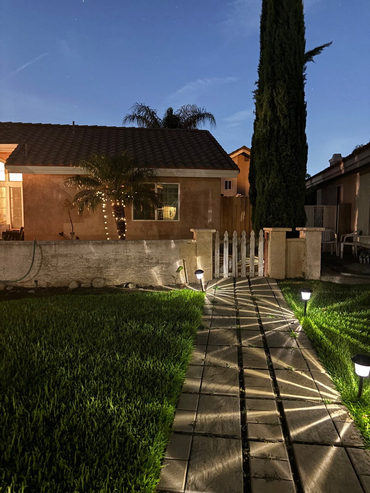 A well-lit pathway leads to the entrance of the property, bordered by freshly maintained grass. The house features a window and is framed by palms and tall greenery. Soft lighting casts shadows across the stone walkway, creating an inviting evening ambiance.