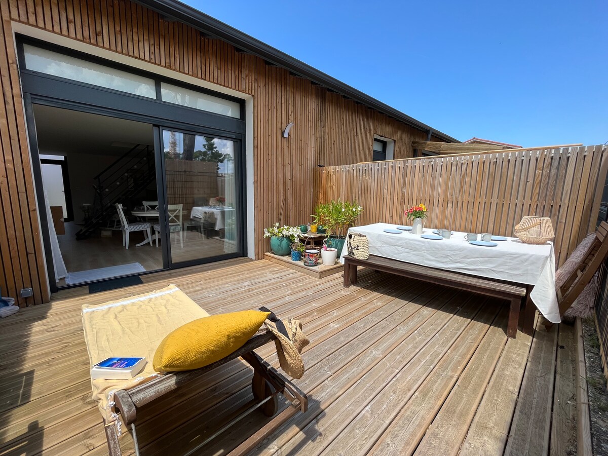 An outdoor terrace features a wooden deck surrounded by potted plants. A large dining table is set for meals, complemented by a sun lounger and a cozy yellow cushion. A glass door provides interior access, allowing natural light to enhance the space.