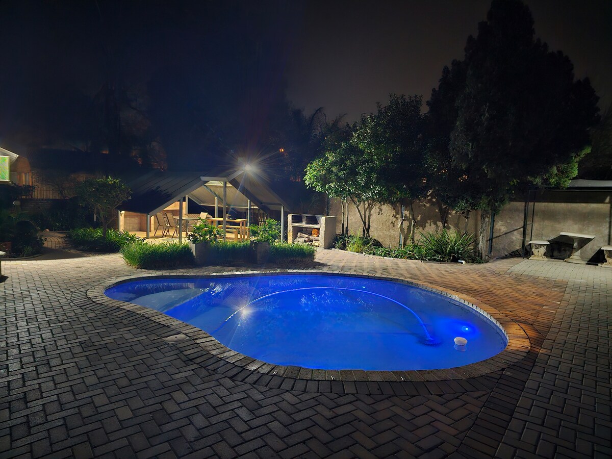 A serene nighttime view of a pool with illuminated turquoise water, surrounded by a brick patio. Lush greenery and outdoor seating are visible in the background, along with a shaded area that includes tables and chairs, creating a tranquil outdoor space.