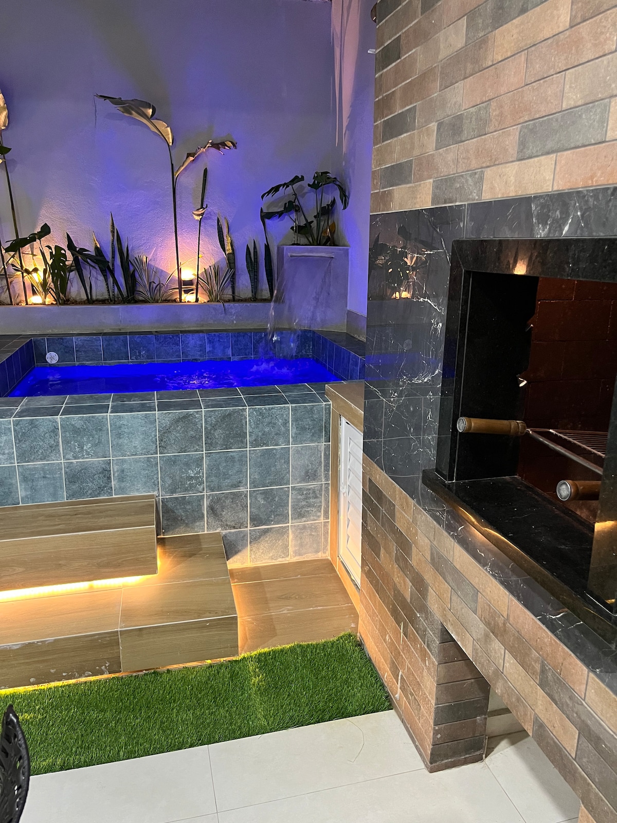 A spa area features a heated hot tub, surrounded by several plants illuminated by soft lighting. A stone wall, complemented by a barbecue grill, adds to the rustic charm of the space. The area is designed for relaxation and social gatherings.