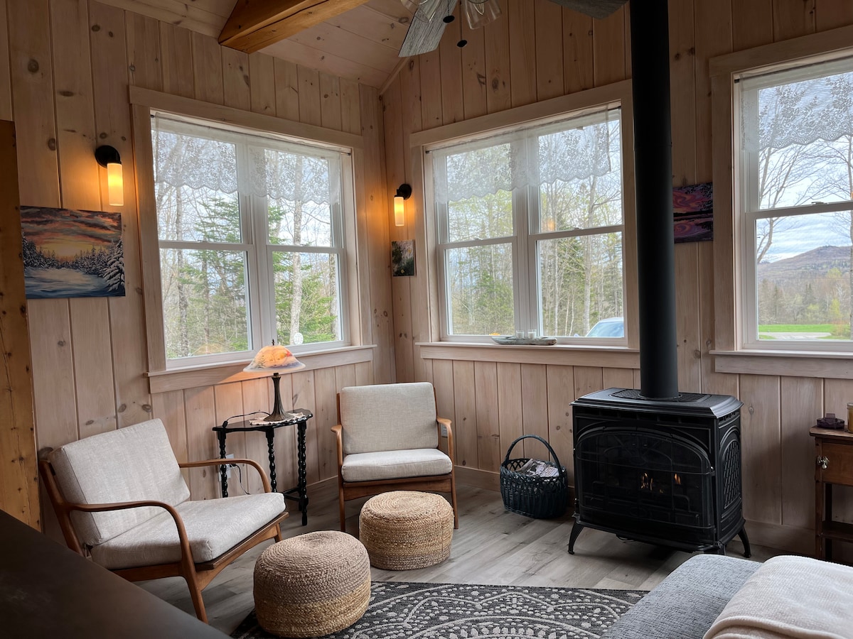 A cozy seating area is framed by large windows, allowing natural light to fill the space. Two upholstered chairs and a round pouf surround a wooden side table. A black stove provides warmth, complemented by subtle wall art and wooden paneling.