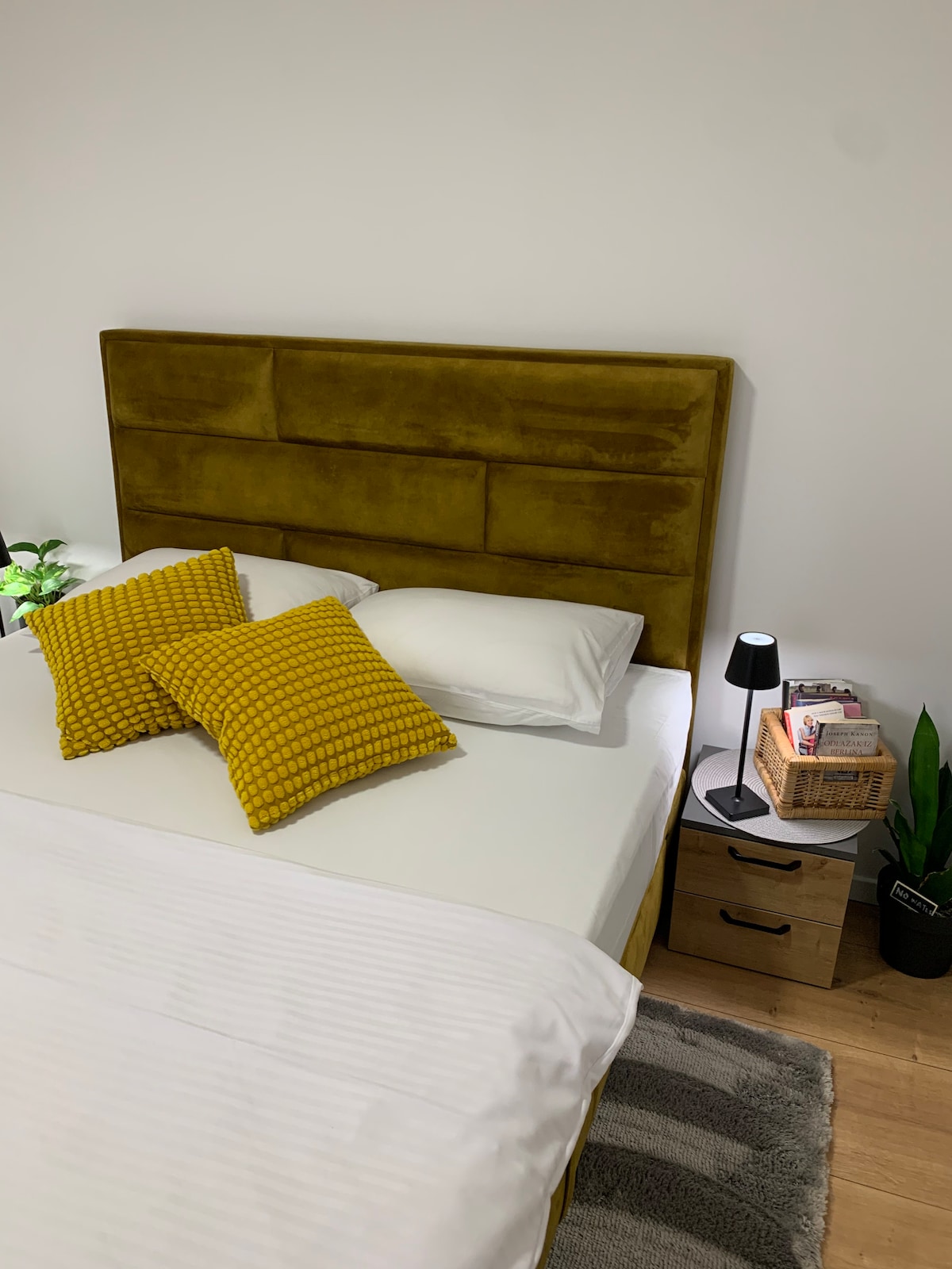 A spacious bed is presented with a plush yellow headboard and soft white linens. Two textured yellow cushions are arranged on the pillow. Adjacent to the bed, a small table holds a decorative lamp and a collection of books, complemented by green plants for a touch of life.