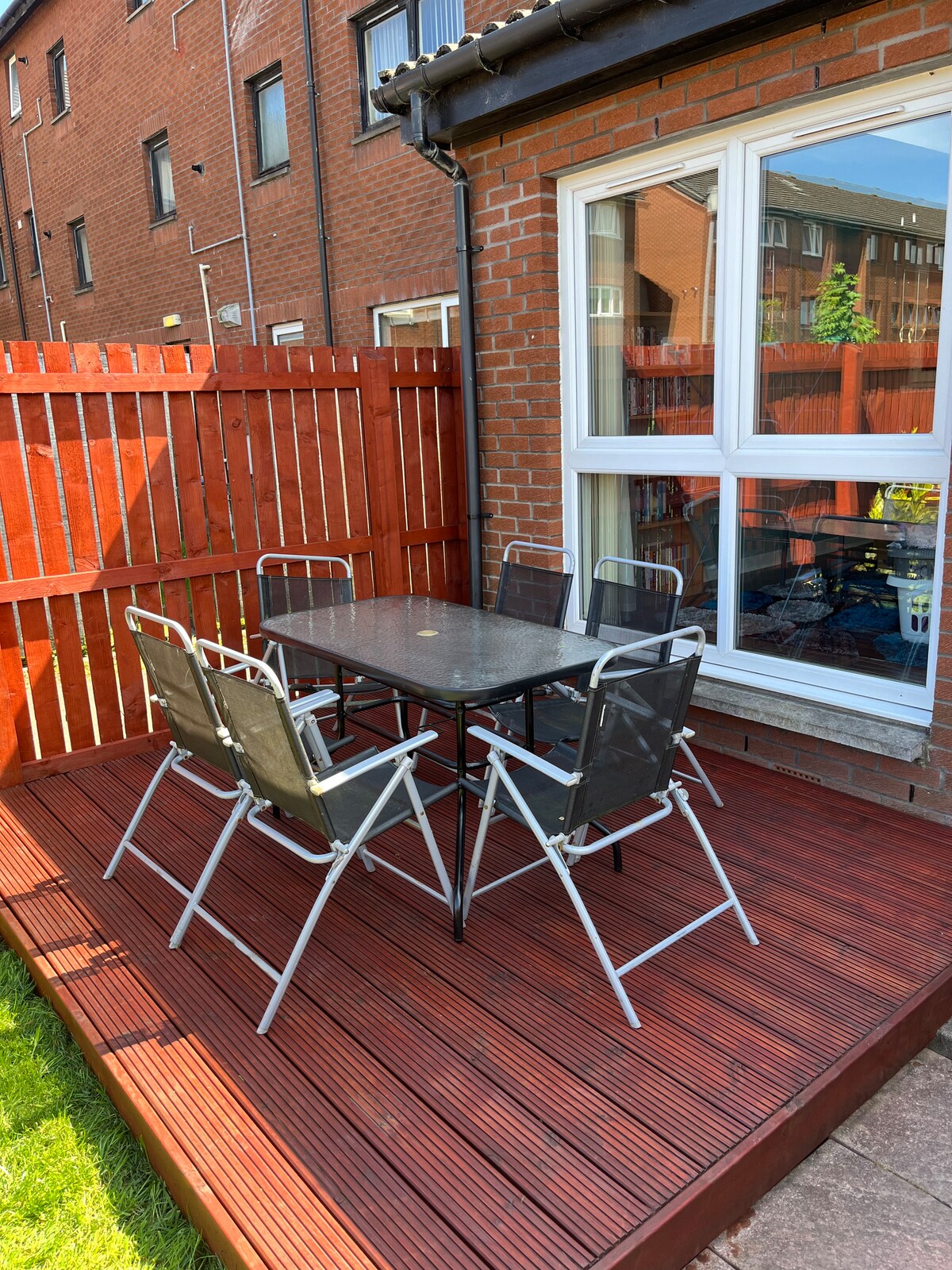 An outdoor seating area features a wooden deck with a table and six metallic chairs, providing a space for al fresco dining. A wooden fence offers privacy, while a well-maintained garden is visible in the background.