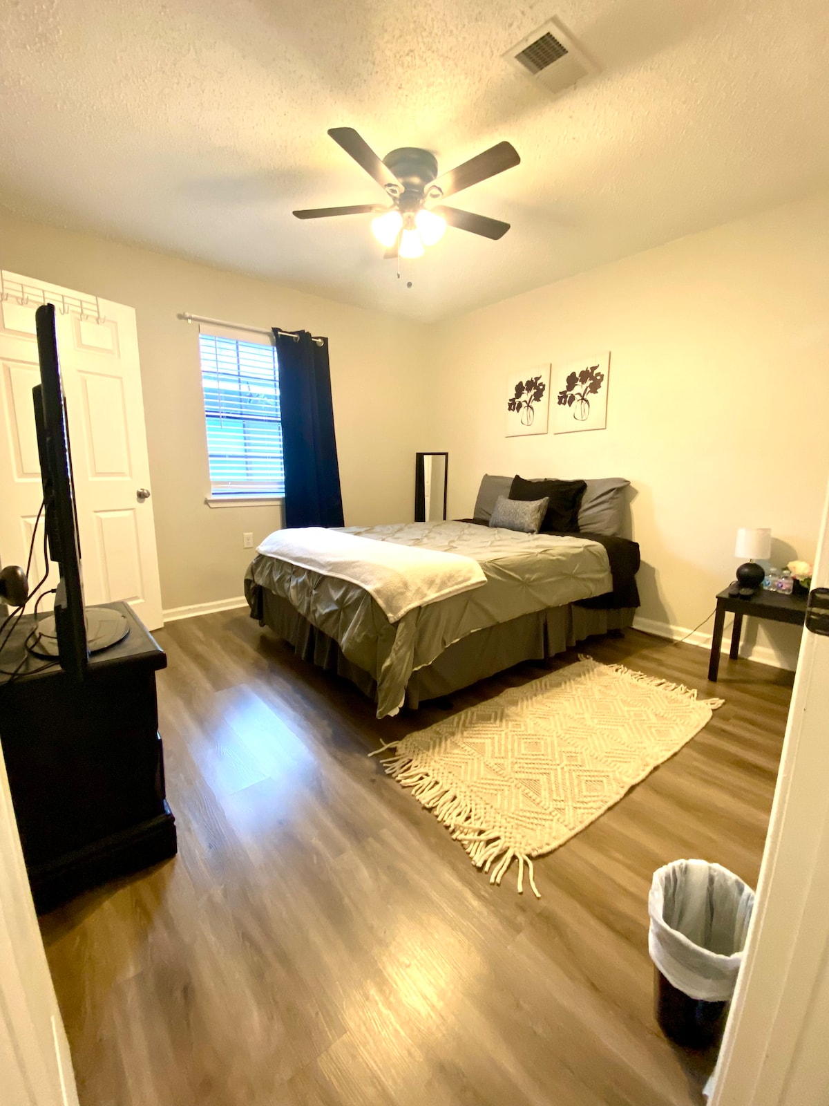 A private room is presented, featuring a spacious bed made with grey bedding. A black dresser stands against one wall, accompanied by a television. A small table with a lamp is situated beside the bed, and a wastebasket is placed in the corner. Natural light enters through a window with blinds.