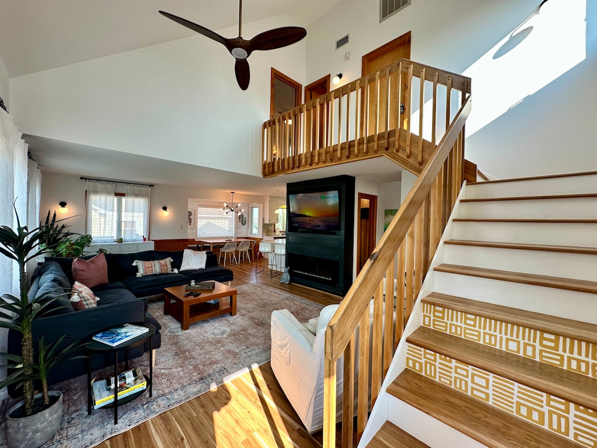 A spacious living area is filled with natural light, showcasing a comfortable sectional sofa and a wooden coffee table. A staircase leads to the upper level, accompanied by a balcony overlooking the living space. Decorative plants are positioned for an inviting touch.