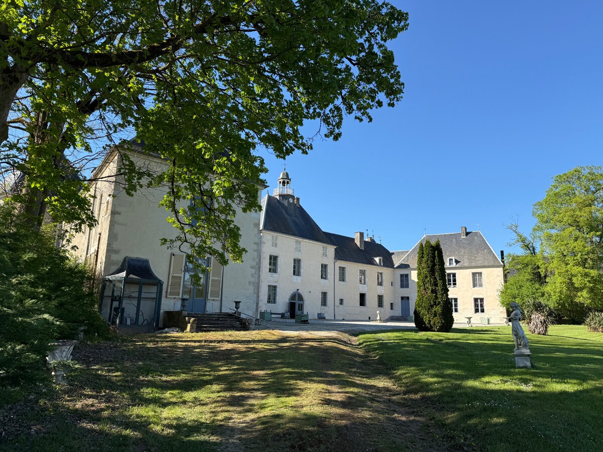 The château is framed by lush greenery, showcasing historical architecture from multiple periods. A clear blue sky provides bright lighting, and the expansive grounds feature manicured lawns and scattered sculptures, offering a serene setting. The façade displays a harmonious structure with a central tower.
