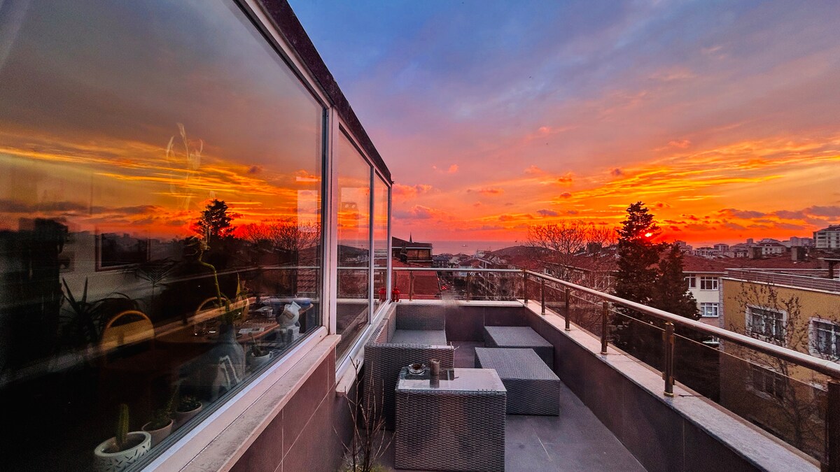 An expansive terrace showcases a stunning sunset, casting warm colors over the sky. Outdoor seating arrangements are visible, while panoramic windows reflect the vibrant hues of the dusk. The space offers a serene setting with a backdrop of trees and buildings.