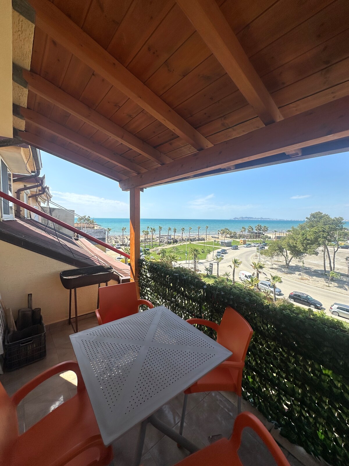 A private balcony features a wooden pergola and a glass-top dining table surrounded by orange chairs. The view includes palm trees lining a road and a glimpse of the sea, creating a serene outdoor space for relaxation.