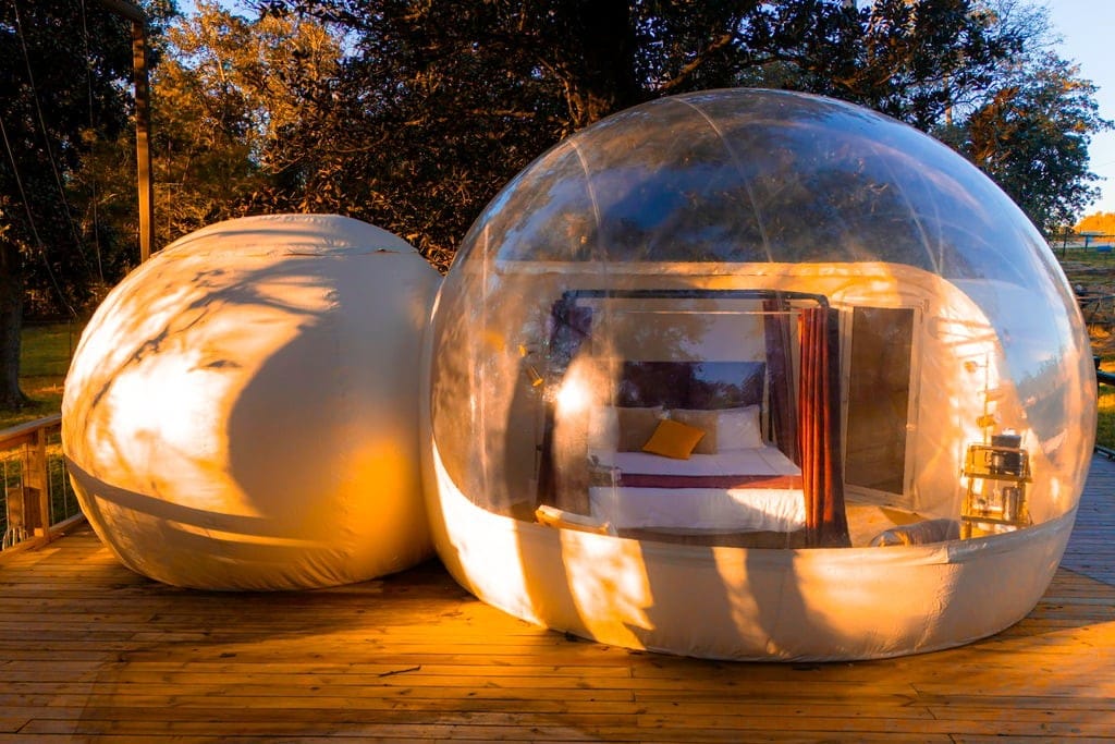 Two inflatable bubbles are positioned on a wooden deck, featuring transparent walls that create an unobstructed view of the surrounding nature. One bubble contains a cozy king bed and is illuminated by soft light, providing a unique sleeping experience under the stars.