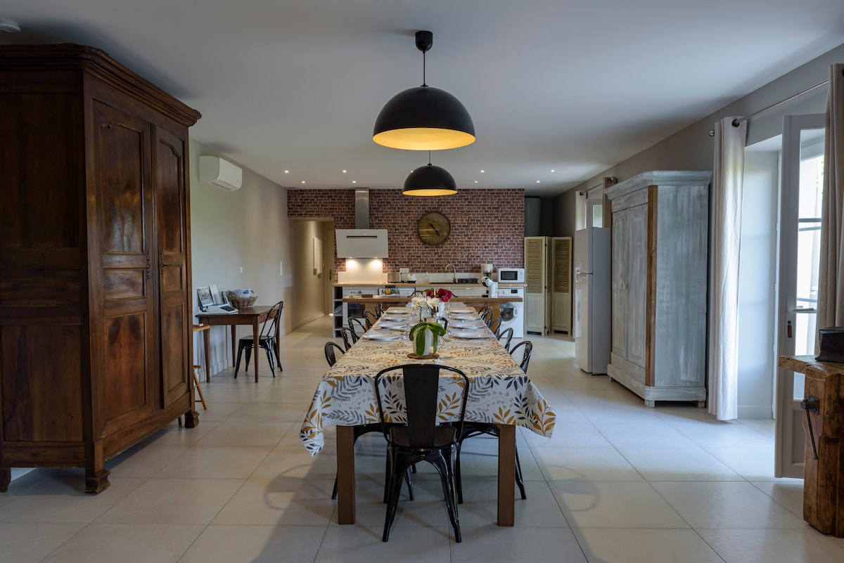 A spacious dining area features a long table set for meals, surrounded by black chairs. A large pendant light illuminates the space, highlighting the textured brick wall. Natural light enters through multiple windows, complemented by elegant curtains.