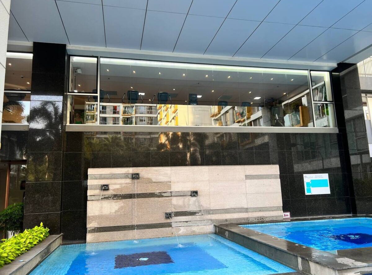 A modern building exterior features a sleek glass facade with a view of a spacious meeting area behind it. Water cascades gently from a wall feature into two tranquil pools below, reflecting the surrounding greenery and architecture.