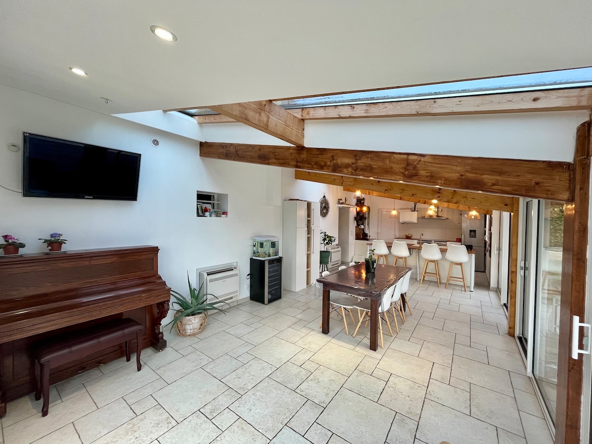 A spacious living area features a grand piano beside a large dining table that comfortably accommodates several guests. Natural light floods the space through a skylight above, illuminating the neutral-toned, tiled floor. The modern kitchen is visible in the background, enhancing the open layout.