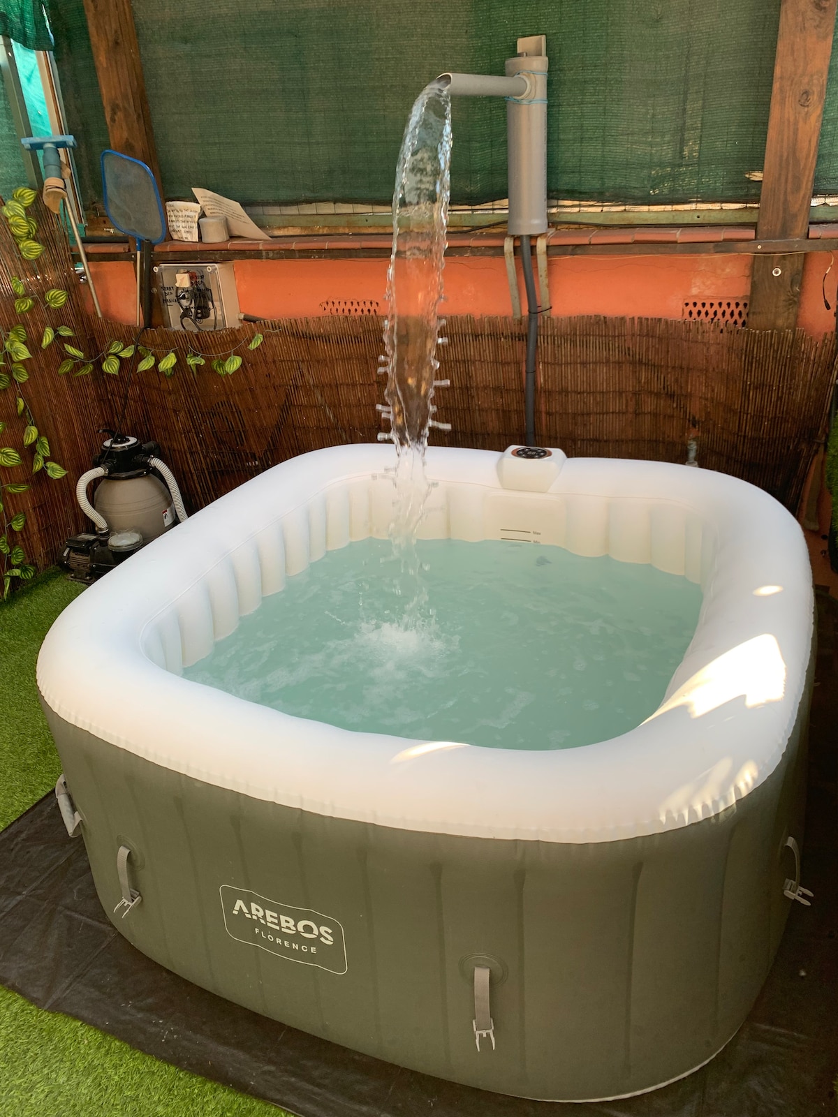 An inflatable jacuzzi is filled with clear water, featuring a cascading fountain effect from a spout. The surrounding area is enhanced with greenery, while a protective covering creates a private oasis. The space is designed for relaxation and leisure.