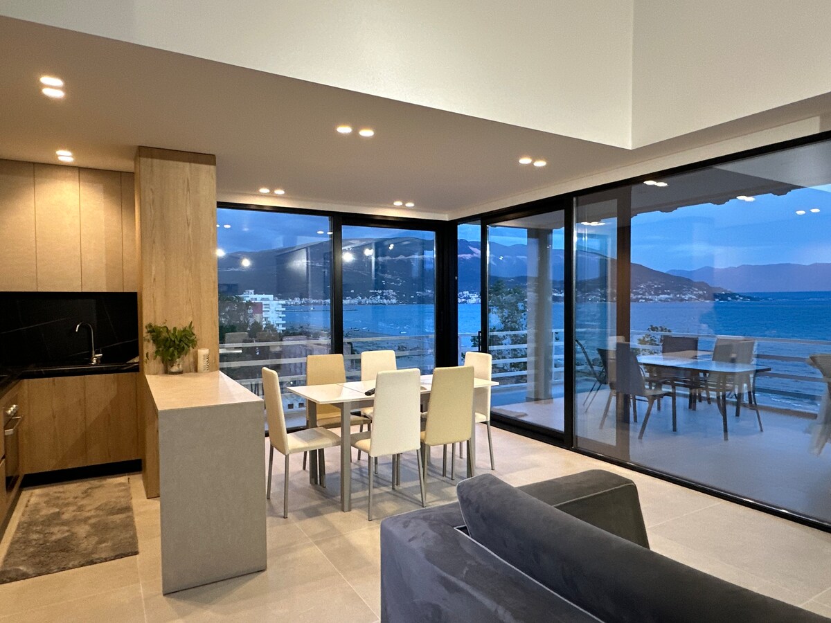 A bright and spacious living area features a modern kitchen and a dining table surrounded by chairs. Expansive glass walls reveal stunning sea views and natural light, while a sofa adds comfort to the room, connecting indoor and outdoor spaces.