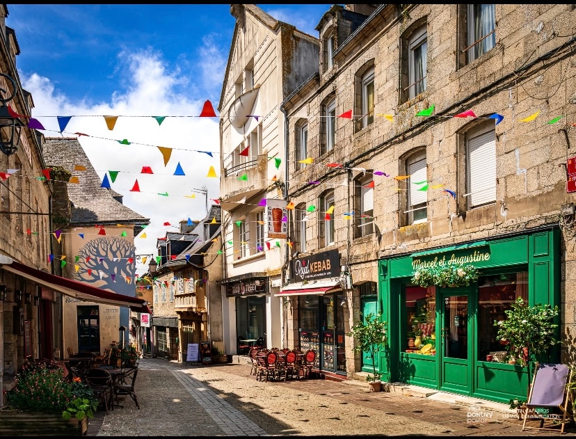 A charming street scene in a town center is illustrated, with colorful bunting strung overhead. Stone buildings with various facades line the street, including a vibrant green storefront. Outdoor seating with tables and chairs is set along the cobblestone path, inviting visitors to relax.