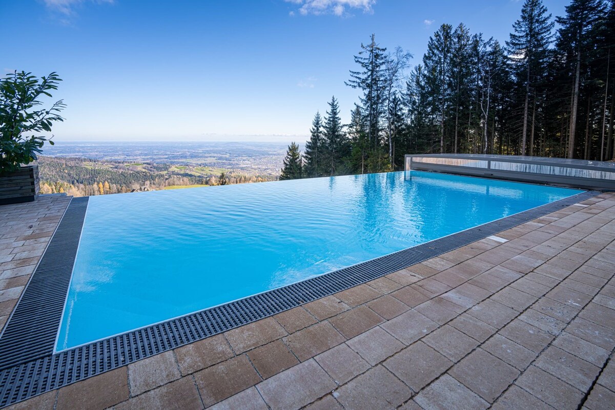 An infinity pool is situated at the edge of a forest, offering panoramic views of the valley below. The water appears calm and inviting, reflecting the clear blue sky, while surrounding trees provide a natural backdrop.