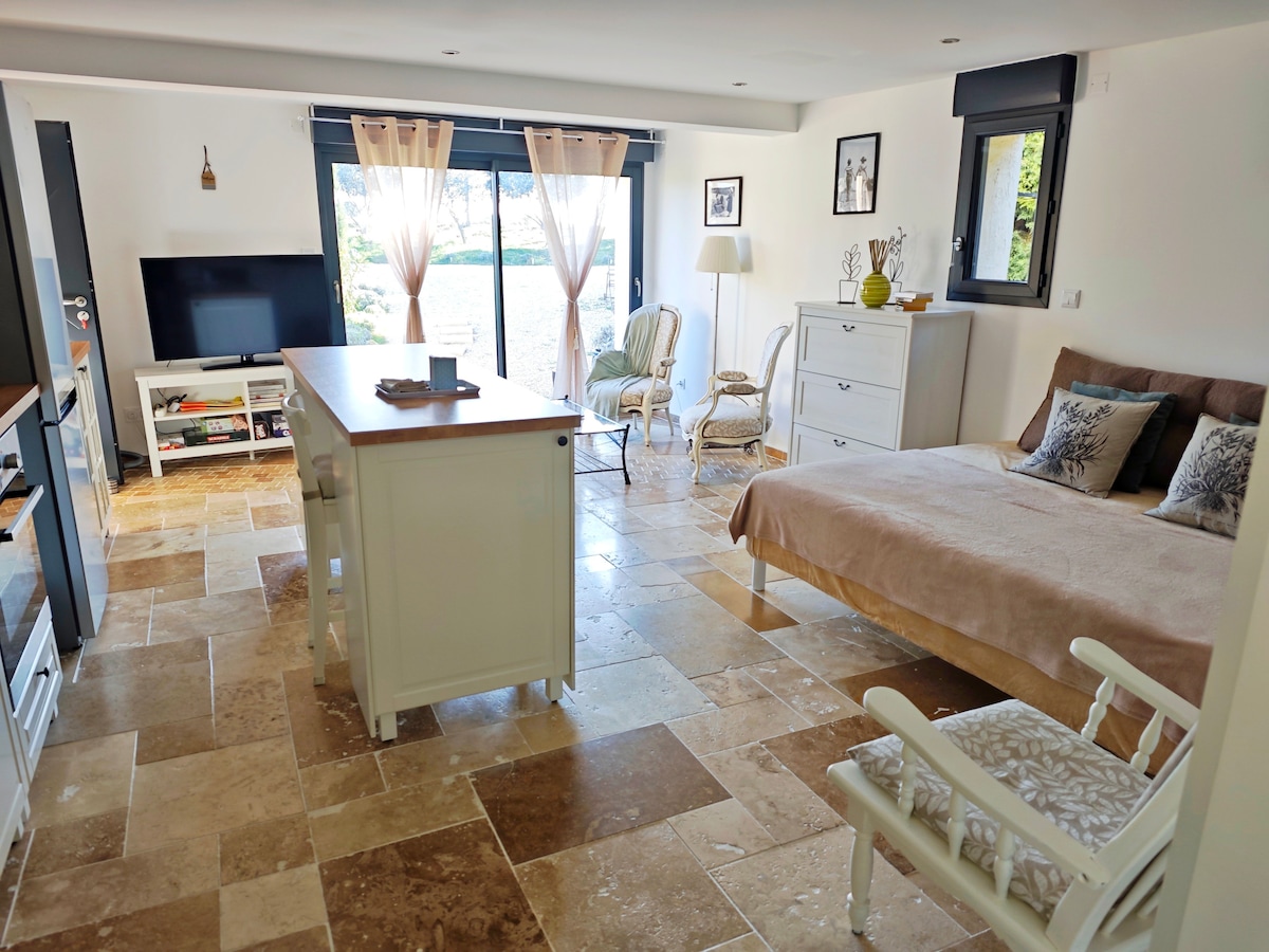 A bright and open living space is showcased, featuring a cozy sleeping area with a spacious bed. Natural light streams through large glass doors, leading to an outdoor view. A small dining table and chairs complement the space, which is finished with elegant stone flooring.