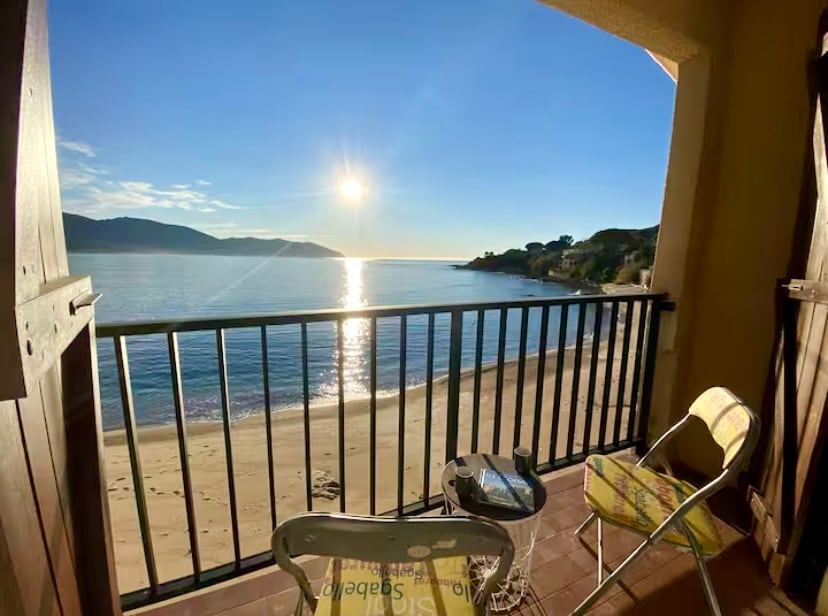 A serene balcony view showcases the calm sea at sunrise, with the sun reflecting off the water. A small table and a folding chair are positioned on the balcony, overlooking the sandy beach and surrounding hills.