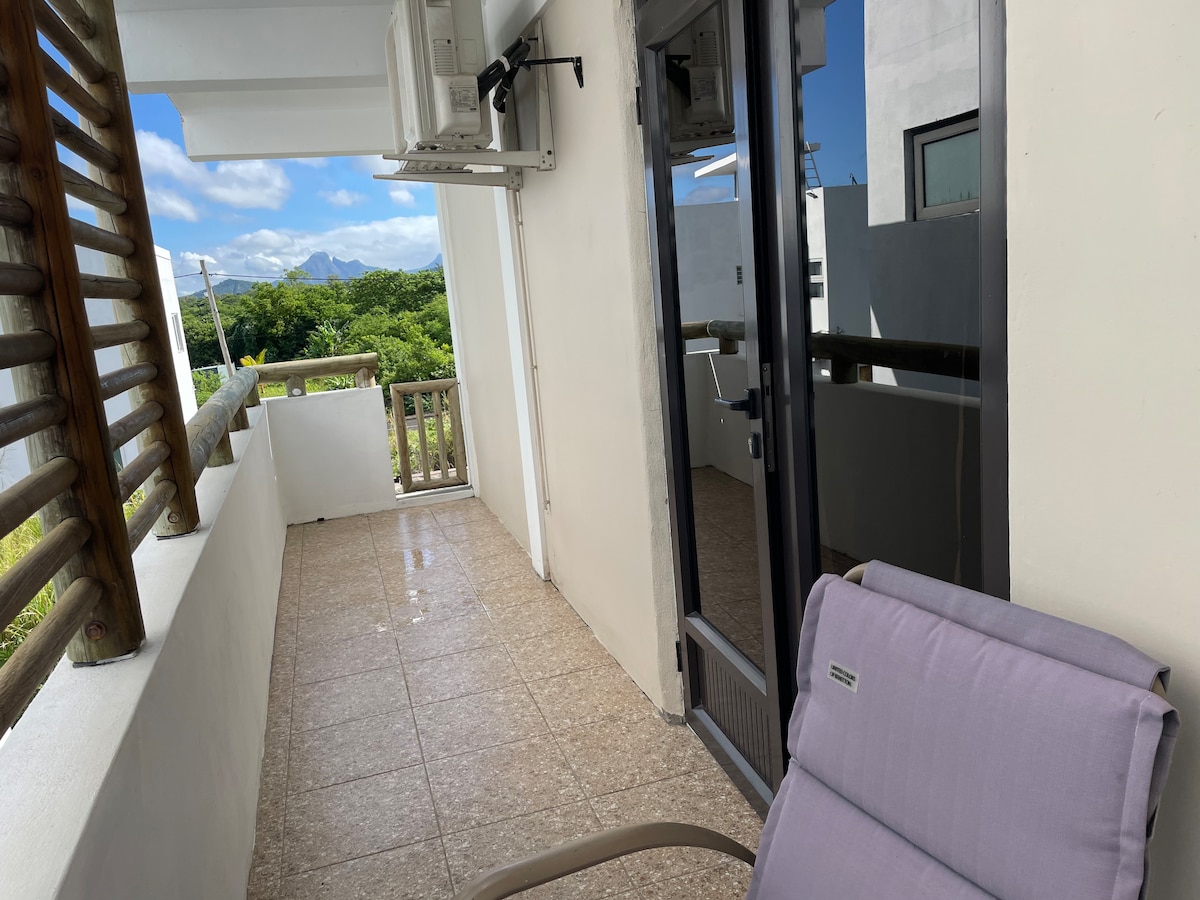 A serene balcony is featured, showcasing tiled flooring with a single chair. Large glass doors provide access to the indoor space, while wooden railings offer an open view of the surrounding greenery and sky. The silhouette of Le Morne Brabant can be seen in the distance.
