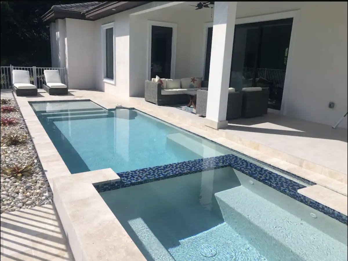 A modern outdoor area showcases a sparkling pool with a shallow entry. Sun loungers rest beside the water, and a covered seating arrangement is seen in the background. Natural stone tiles frame the pool, while decorative pebbles and plants enhance the area.