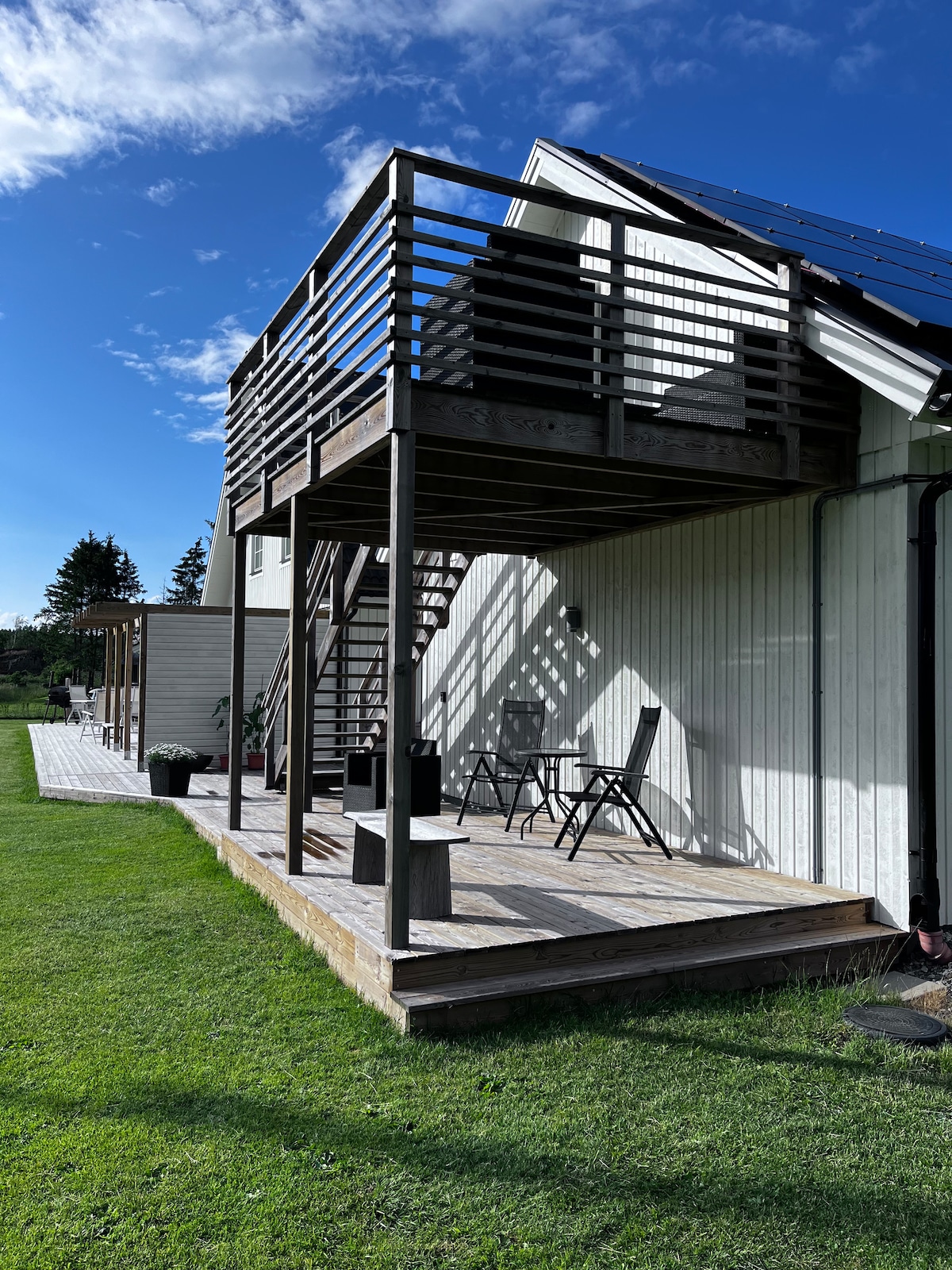 A deck is visible on the upper level of a white, two-story building. A table and two chairs are positioned on the deck, providing a space for outdoor relaxation. The surrounding lawn is well-kept, and the sky is clear with scattered clouds.