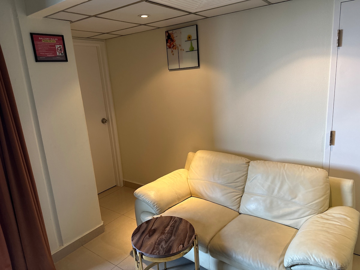 A light-colored leather sofa is positioned next to a small round table with a wooden top. A framed piece of artwork is displayed on the wall above the sofa. Soft lighting is provided by overhead fixtures, contributing to a serene environment.