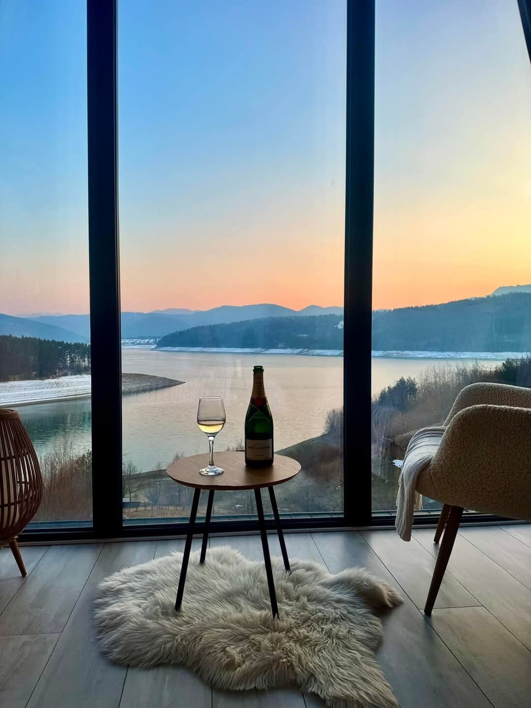 A serene view is captured through a large window, showcasing a lakeside landscape at sunset. A small table is set with a glass of champagne and a bottle, while two modern chairs are positioned nearby. The reflection of soft colors in the water enhances the ambiance.