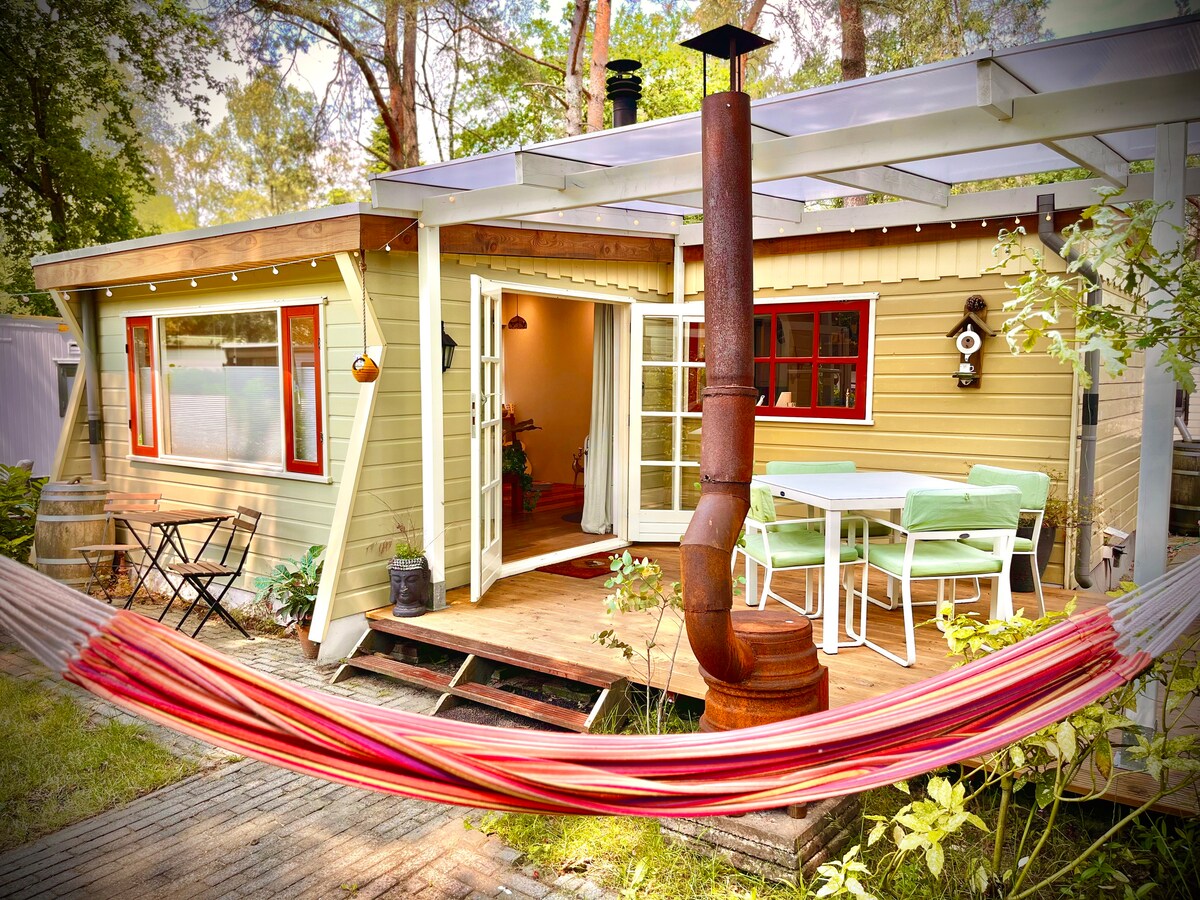 The exterior of the chalet features a welcoming porch with seating for four, surrounded by nature. A vibrant hammock is suspended nearby, inviting relaxation. A rustic wood-burning stove adds character, while large windows provide ample natural light, blending indoor and outdoor spaces seamlessly.