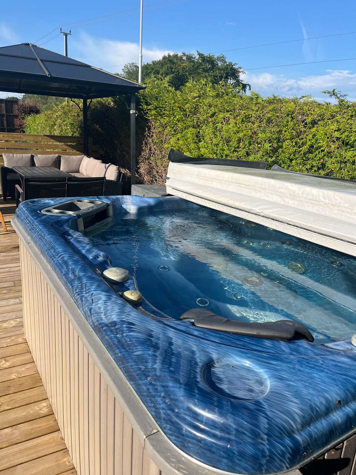 A hot tub is positioned on a wooden deck, featuring a protective cover. Surrounding the tub, there is comfortable outdoor seating arranged beneath a canopy, with greenery providing privacy and a tranquil atmosphere.