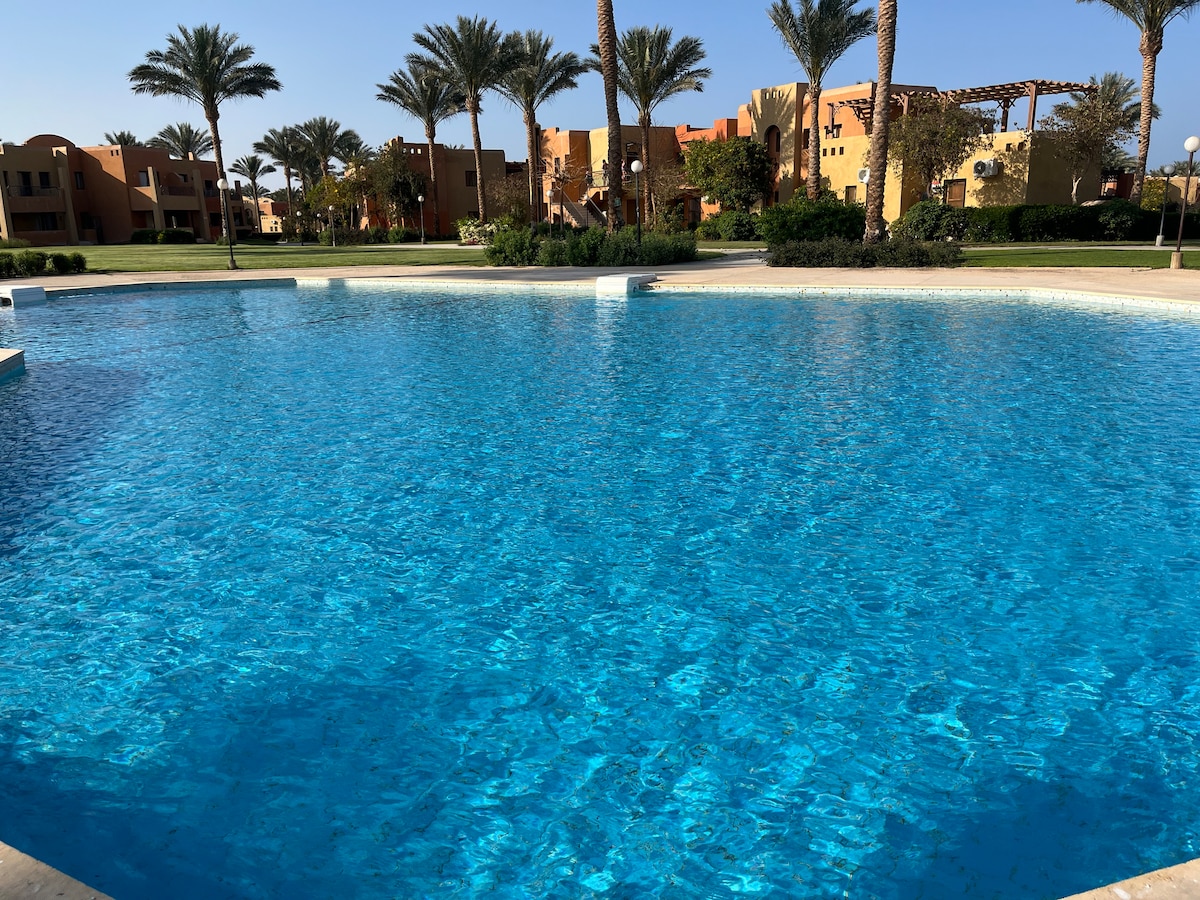 A large swimming pool is surrounded by palm trees, with sunlight reflecting off the clear blue water. In the background, low-rise buildings in warm tones can be seen among the landscaped grounds.