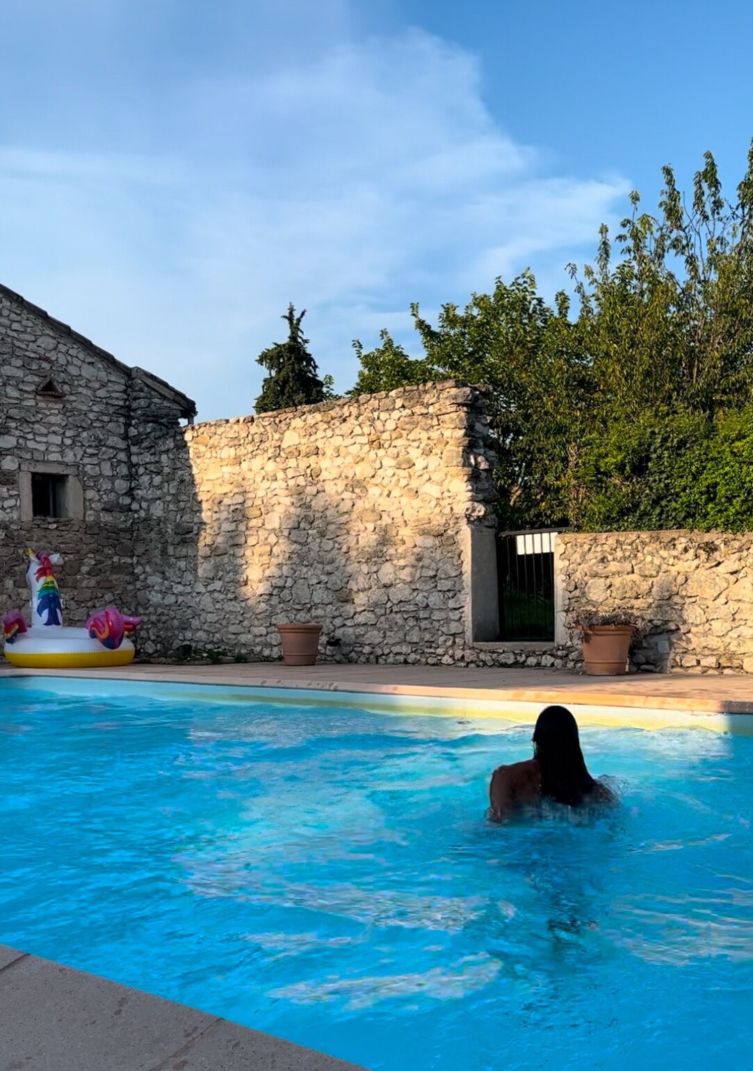 A shared pool is surrounded by rustic stone walls, offering a peaceful atmosphere. A person is seen swimming in the crystal-clear water. Lush greenery is visible in the background, contributing to the serene setting, while a colorful inflatable float adds a playful touch.