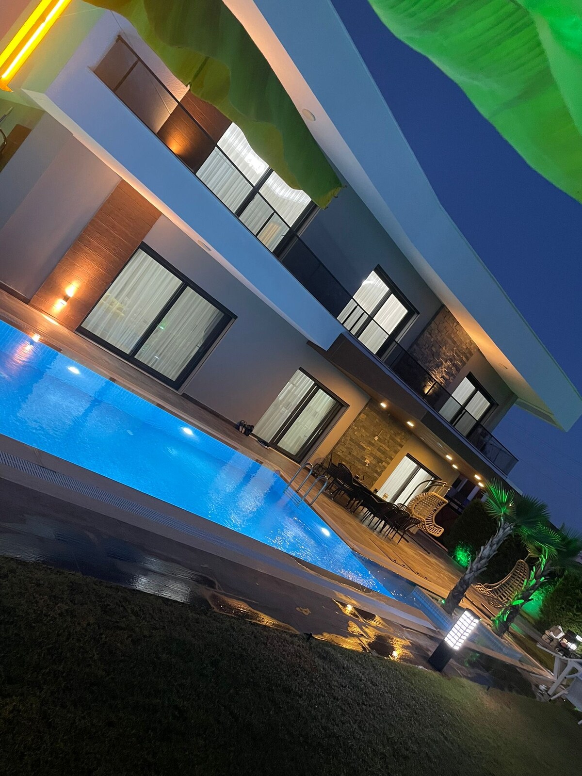 A modern villa is showcased from the side, featuring a shimmering pool that reflects the evening lights. Large glass doors and windows provide visibility into the indoor spaces, while outdoor seating is arranged near the pool. Lush greenery surrounds the area, enhancing the serene environment.