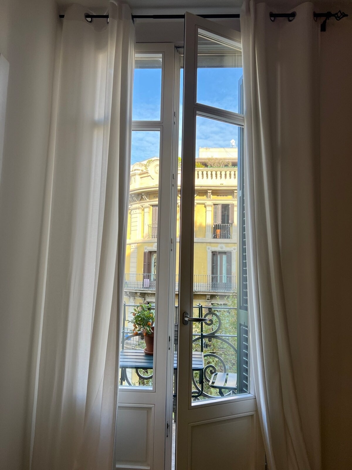 Tall windows with white curtains frame a view of a neighboring building, showcasing ornate architecture and a small balcony with potted plants. Soft natural light enters the space, enhancing the airy atmosphere.