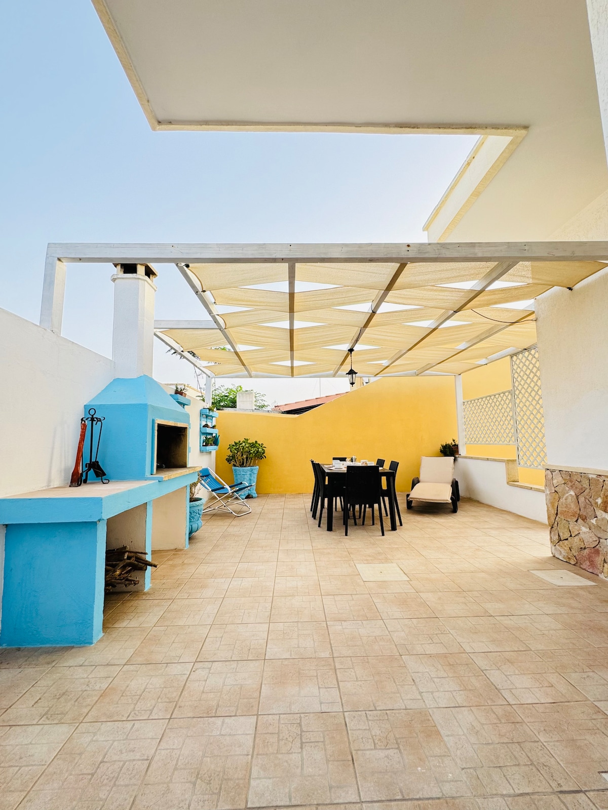 An outdoor patio area is presented, featuring a dining table with chairs beneath a removable canopy. A barbecue station is located to one side, with blue accents adding a touch of color. Potted plants line the space, providing a welcoming environment.